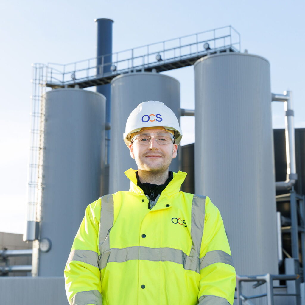 A person wearing a white hard hat and bright yellow safety jacket stands in front of large industrial tanks and pipes at a facility.