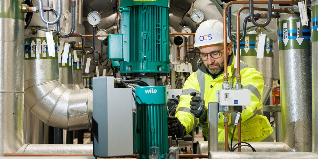 A worker in a hard hat and high-visibility jacket adjusts equipment among pipes and machinery in an industrial facility, surrounded by gauges and green pumps.