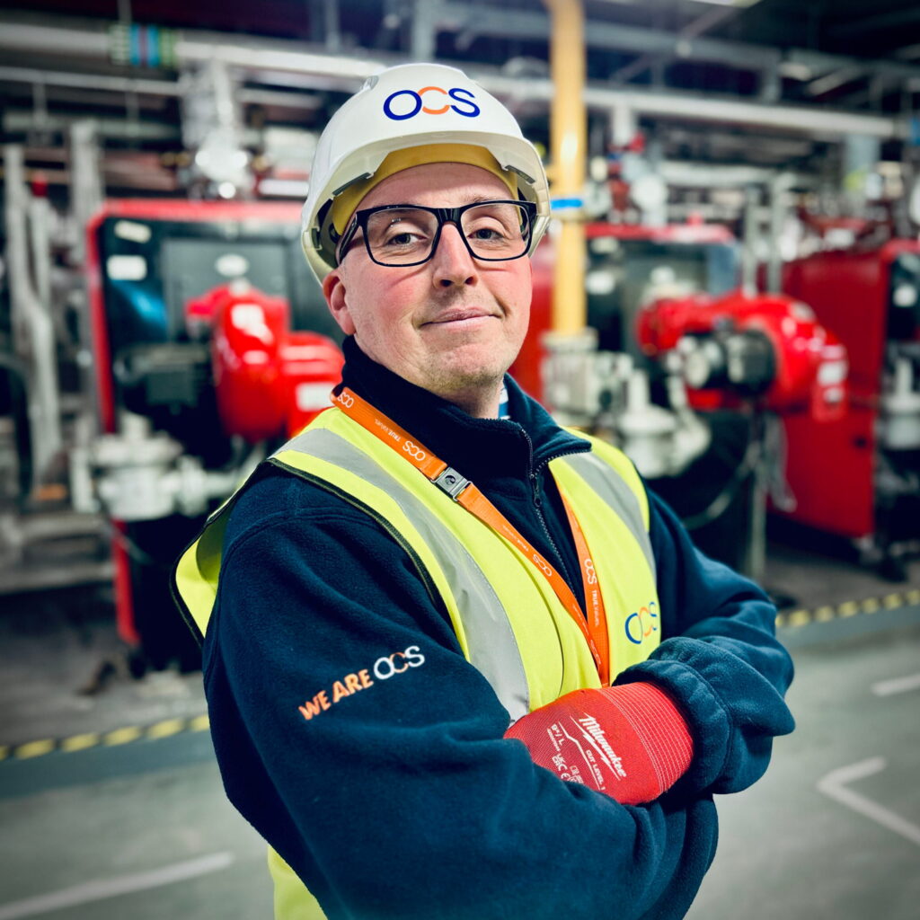A confident worker wearing a hard hat, safety glasses, and a high-visibility vest stands with folded arms in an industrial setting, with machinery and safety lines visible in the background.