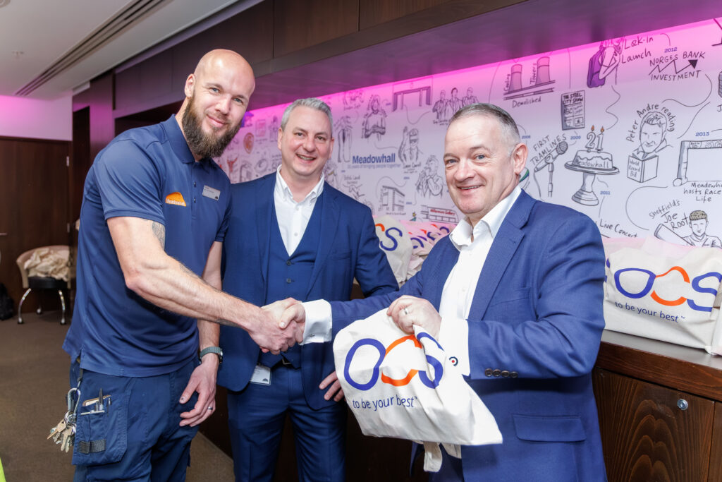 A man in a blue polo shirt shakes hands with a man in a suit whilst another man in a suit stands beside them, all smiling. They hold bags with a colourful “OQS” logo in front of a wall with illustrated drawings.