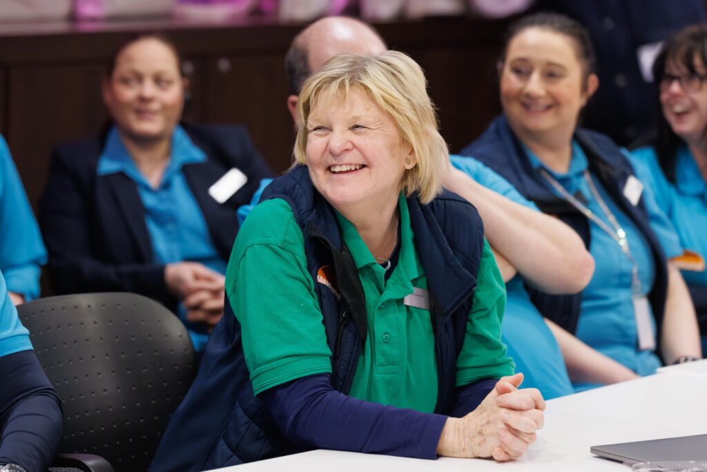 A group of people in uniforms sit at a table, smiling and laughing. The focus is on a woman in a green shirt and blue waistcoat who is sitting at the front, appearing happy and engaged.
