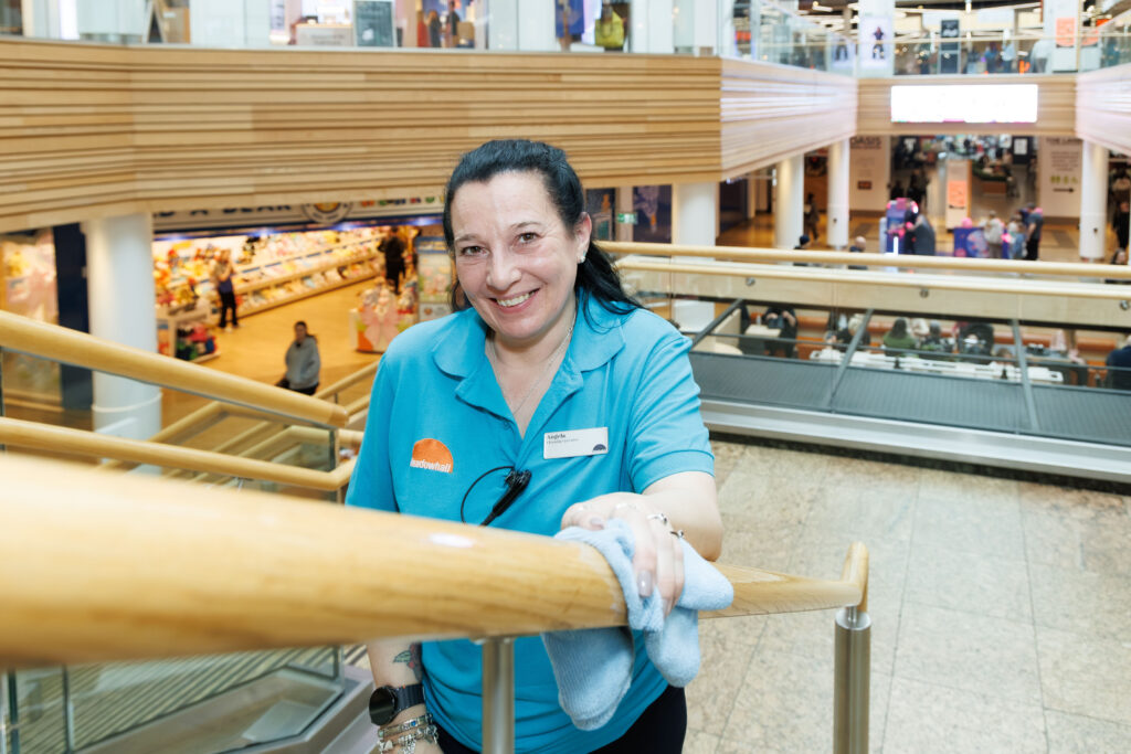 A smiling woman in a teal uniform wipes a wooden handrail in a shopping centre, with shops and shoppers visible in the background.