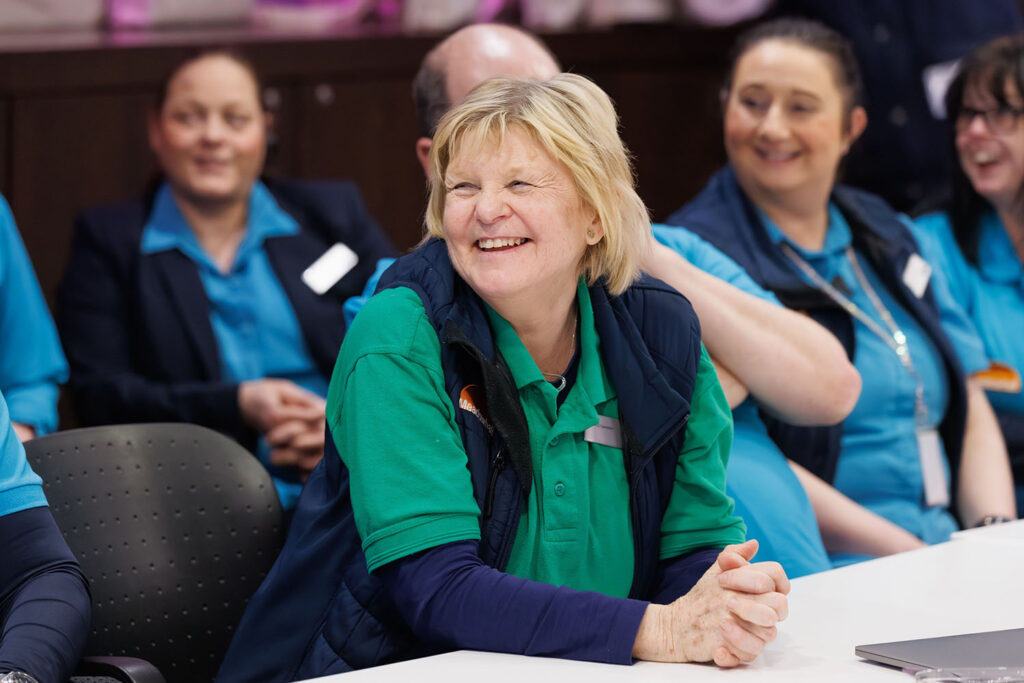 A group of people in uniforms sit together at a table, smiling and laughing. The focus is on a woman in a green shirt and blue vest who is smiling brightly. Others around her also appear happy and engaged.