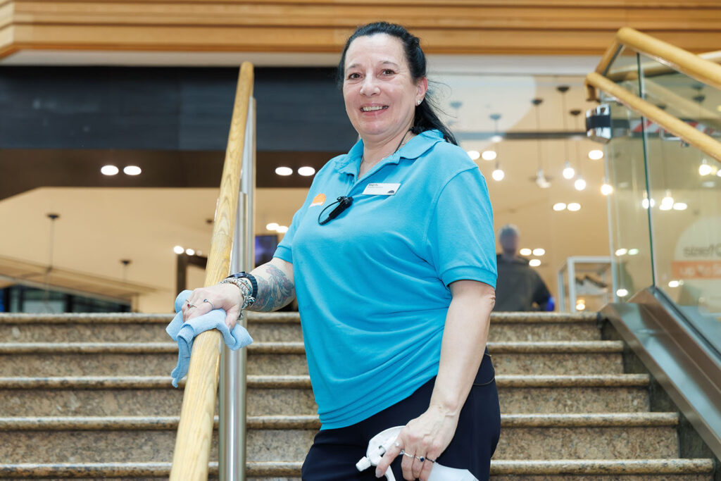 A woman in a blue polo shirt smiles while cleaning a stair railing in a well-lit indoor space, holding a cloth and spray bottle.