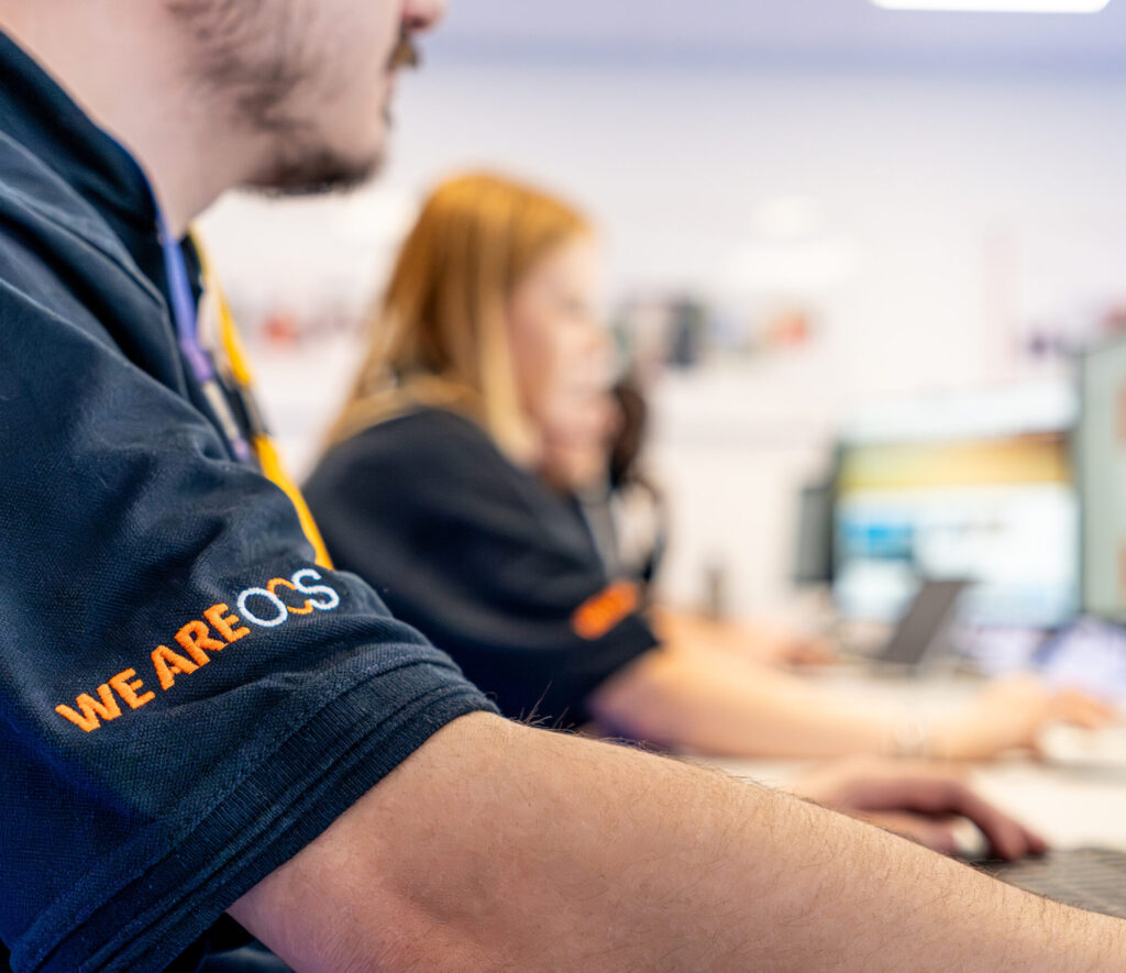 A close-up of a person working at a computer in an office, wearing a navy shirt with WE ARE COS on the sleeve. Other people are also working at computer stations in the blurred background.