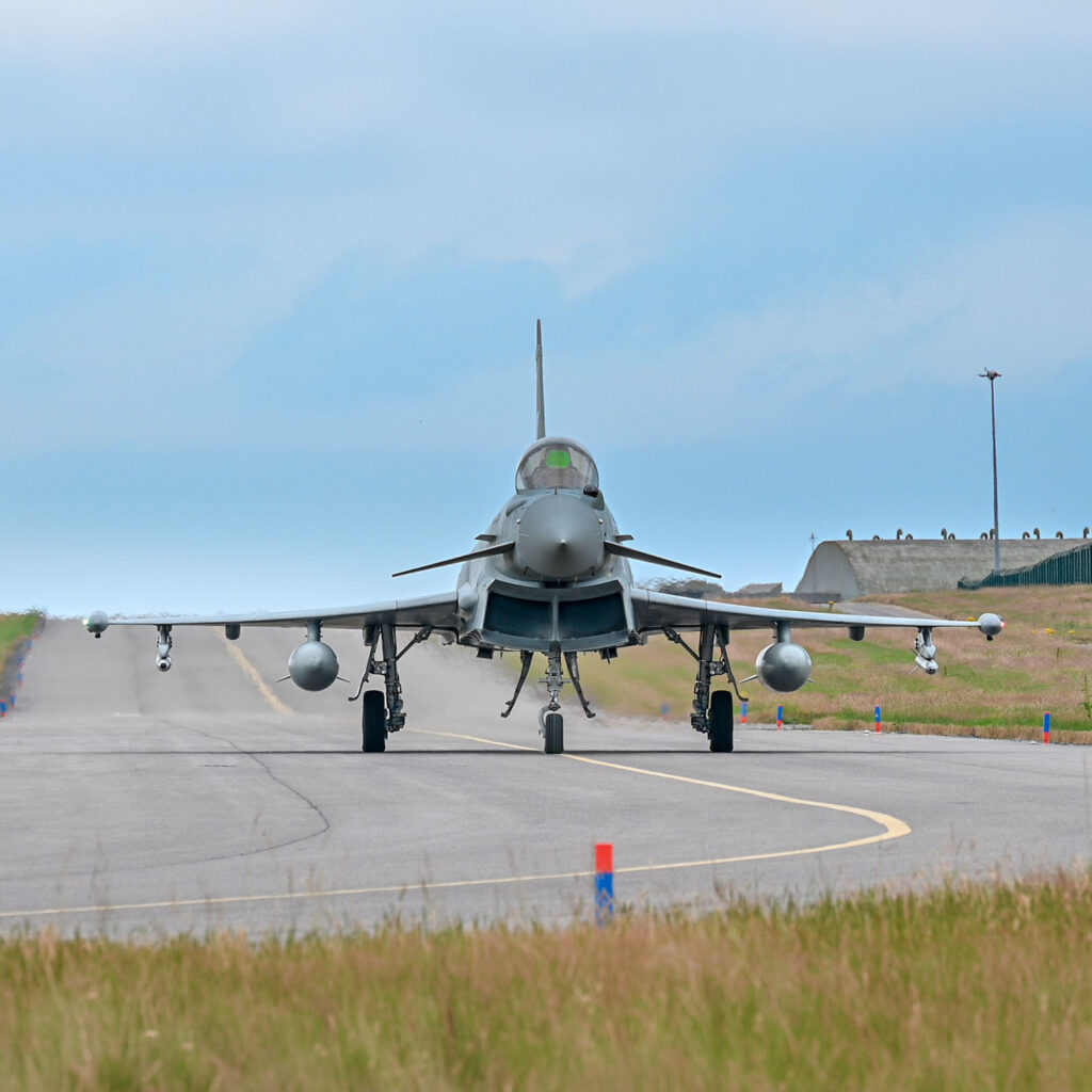 A military fighter jet seen from the front sits on a runway, ready for take-off, with grassy areas on either side and a hangar visible in the background under a partly cloudy sky.