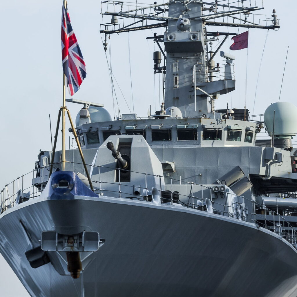 Close-up view of the bow of a naval warship with various aerials, radar equipment, and the British naval ensign flag flying at the stern. The ship appears modern and is shown against a pale sky.