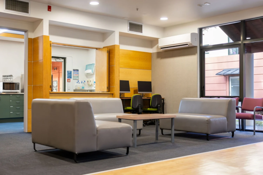 A modern lounge area with grey sofas, a wooden coffee table, and a nearby workspace featuring computers and chairs. The space is well-lit with natural and artificial light.