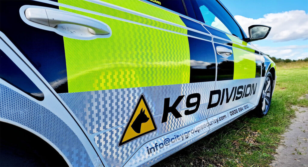 Side view of a black and yellow police car labelled K9 DIVISION with a dog head symbol, parked on grass under a blue sky with clouds. Contact info and a website are visible on the car door.