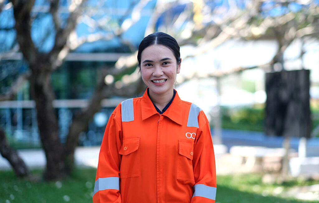 A woman wearing an orange safety uniform with reflective strips stands outdoors, smiling at the camera. There are trees and a building with glass windows in the blurred background.