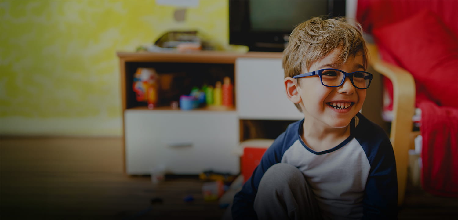 A young boy with glasses smiles and sits on the floor in a cosy, colourful room with toys and art supplies on a shelf behind him.