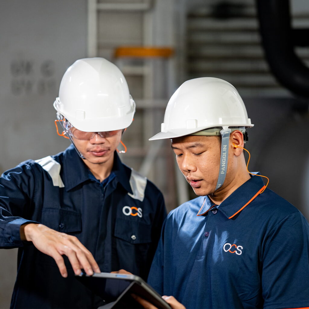Two workers wearing white safety helmets and dark uniforms look at a tablet together in an industrial setting, discussing something with focus.