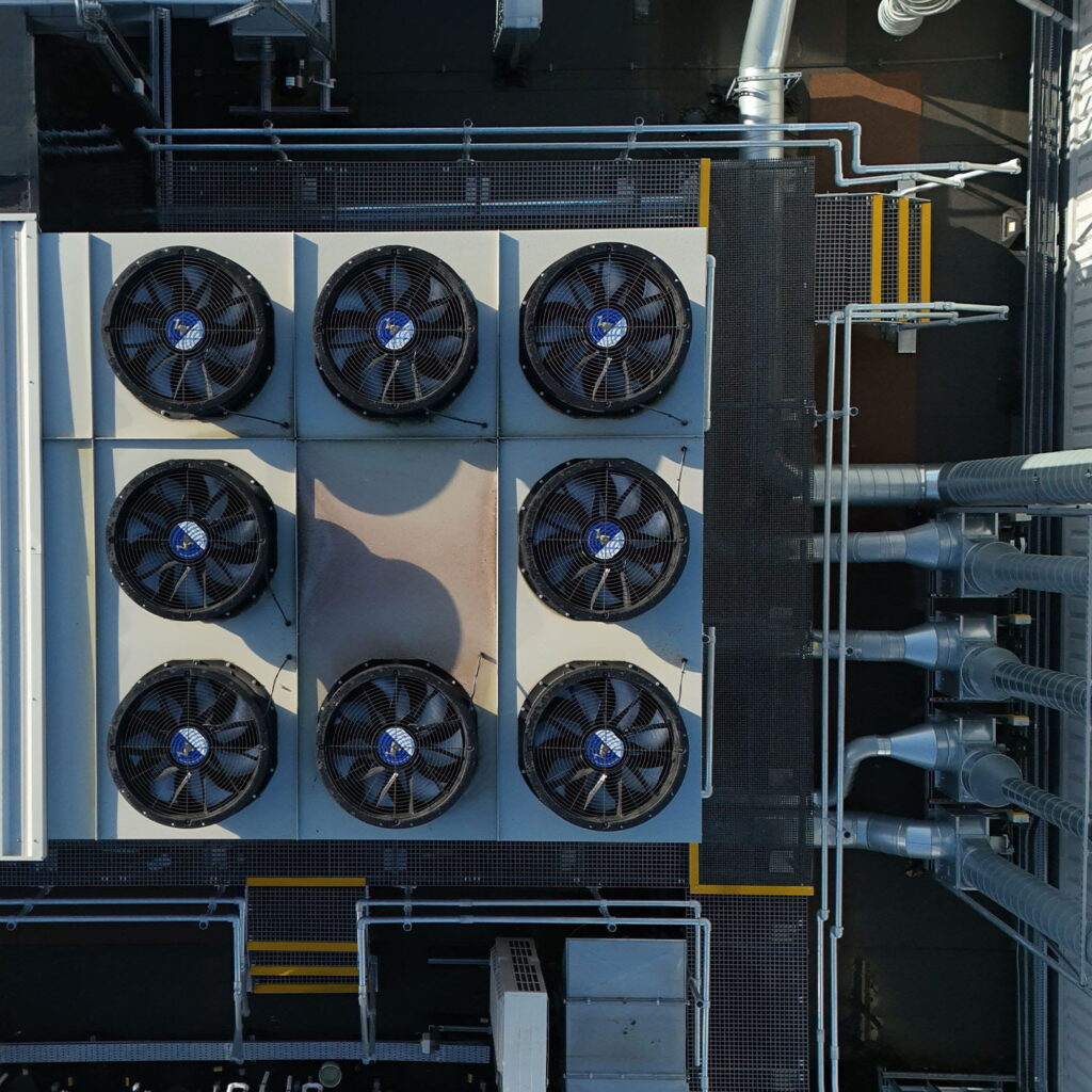 Aerial view of eight large industrial ventilation fans arranged in two rows on a rooftop, surrounded by pipes, metal gratings, and walkways.