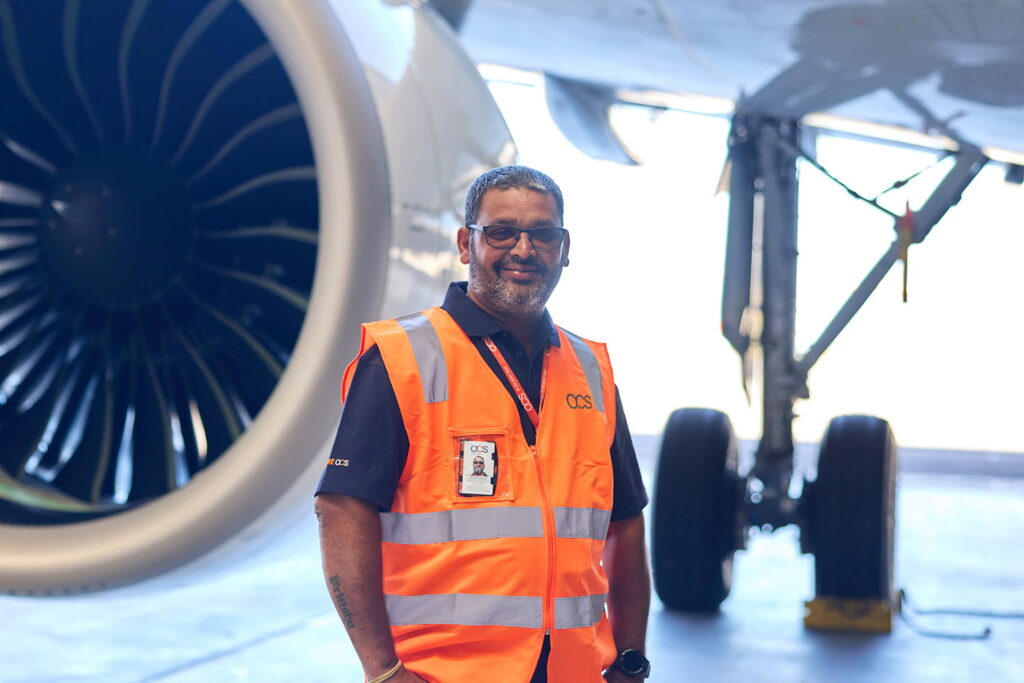 A man wearing glasses, a bright orange safety vest, and an ID badge stands smiling in front of a large airplane engine inside a hangar. The aircraft’s landing gear is visible in the background.