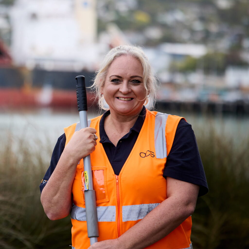 A woman wearing an orange safety vest and holding a mop stands outdoors near water, with a blurred ship and greenery in the background, smiling at the camera.