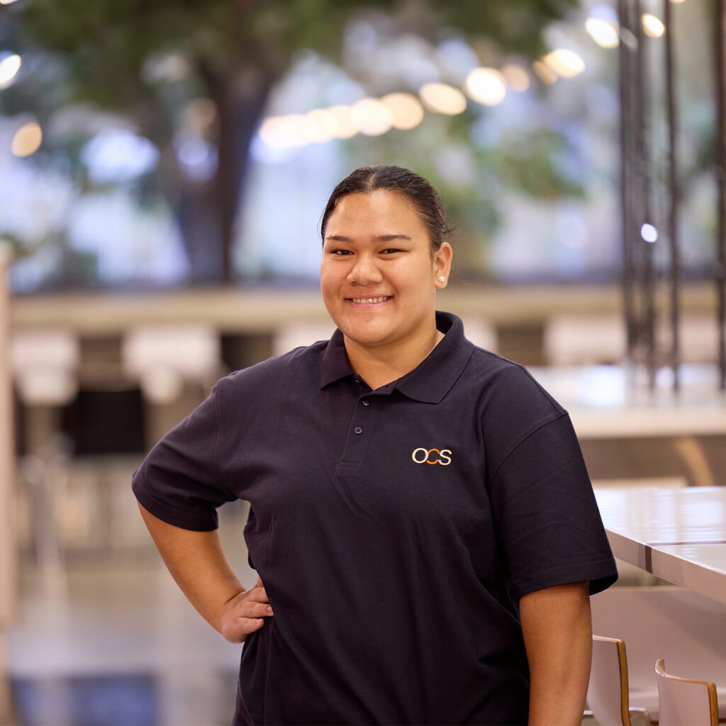 A person wearing a navy blue OCS polo shirt stands indoors, smiling with one hand on their hip. The background is softly blurred, showing tables, chairs, and greenery outside.