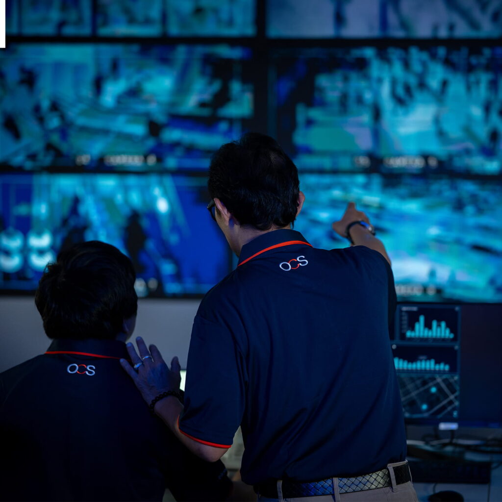 Two people in dark uniforms monitor multiple security screens, with one person pointing at the surveillance footage. The room is dimly lit and filled with blue light from the monitors.