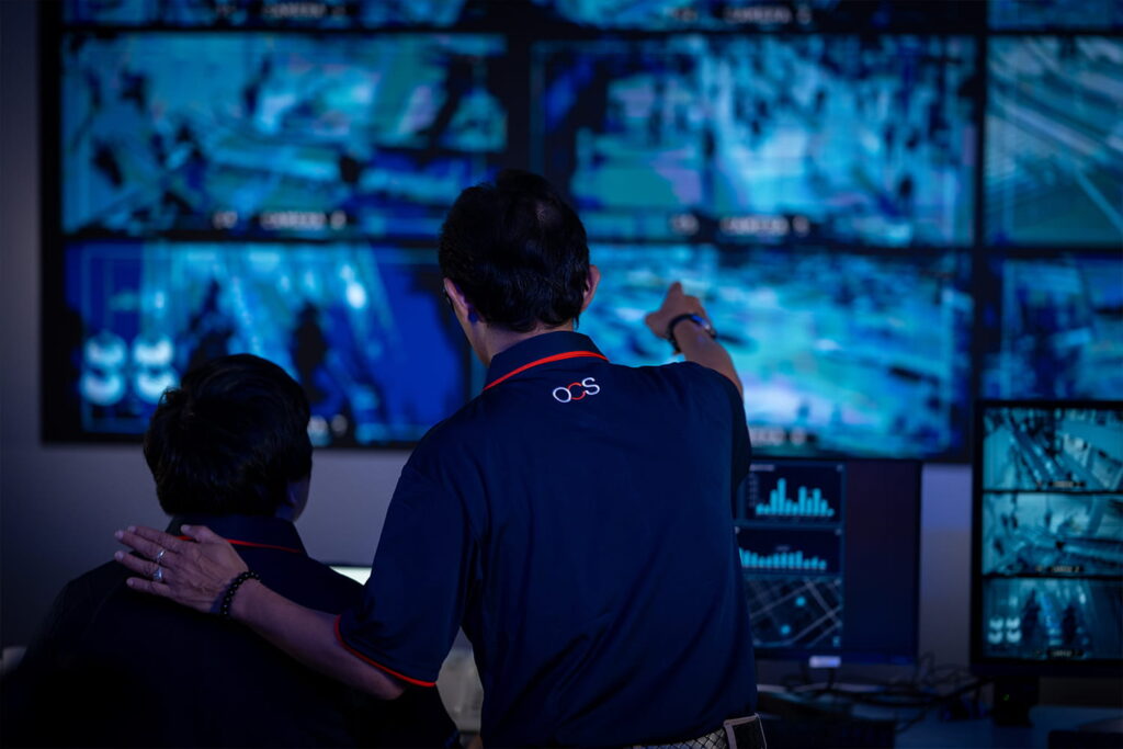 Two people in a dark room monitor multiple surveillance screens; one person is pointing at the screens while the other looks on. Both wear matching dark shirts with “OCS” written on the back.