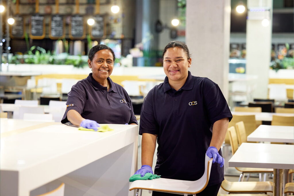 Two women in dark blue uniforms and purple gloves are cleaning tables and chairs in a brightly lit cafeteria. Both are smiling, holding yellow cloths, and appear to be tidying the area.