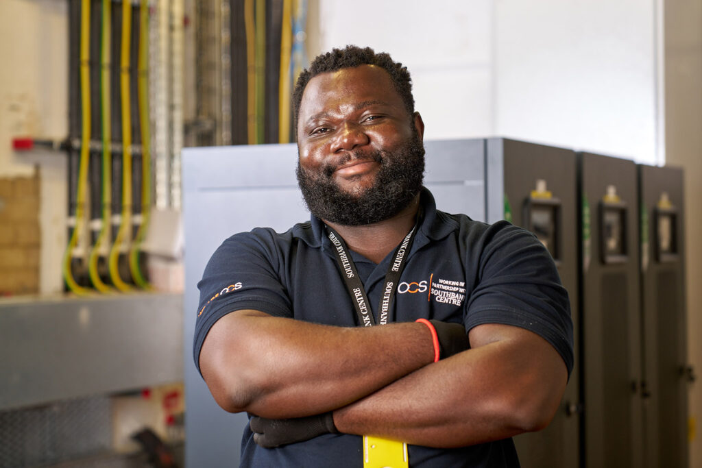 A man with a beard stands with arms crossed, smiling confidently in a technical room with servers and cables in the background. He wears a dark polo shirt and a lanyard.
