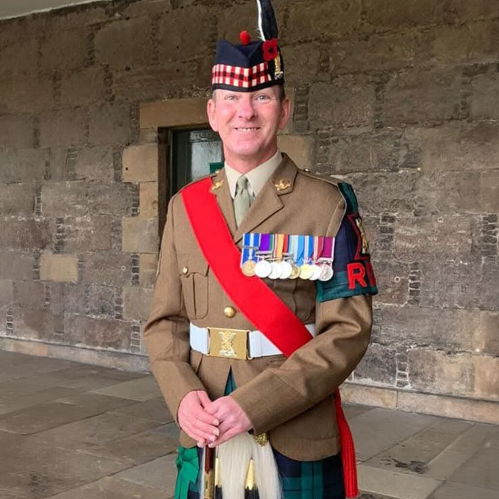 A smiling man in a brown military uniform with a red sash stands in front of a stone wall. He is wearing a kilt, a hat with a feather, and several medals on his chest.