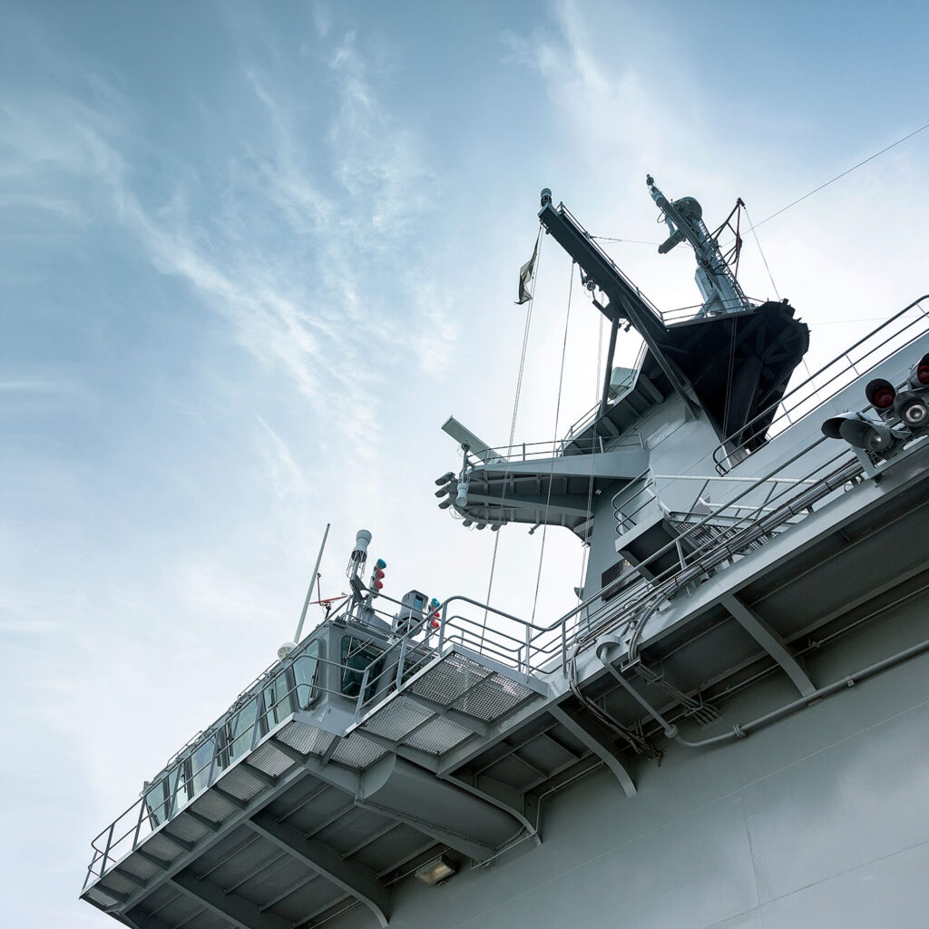 The image shows the upper deck and control tower of a large naval ship, possibly an aircraft carrier, with aerials and equipment against a partly cloudy sky.