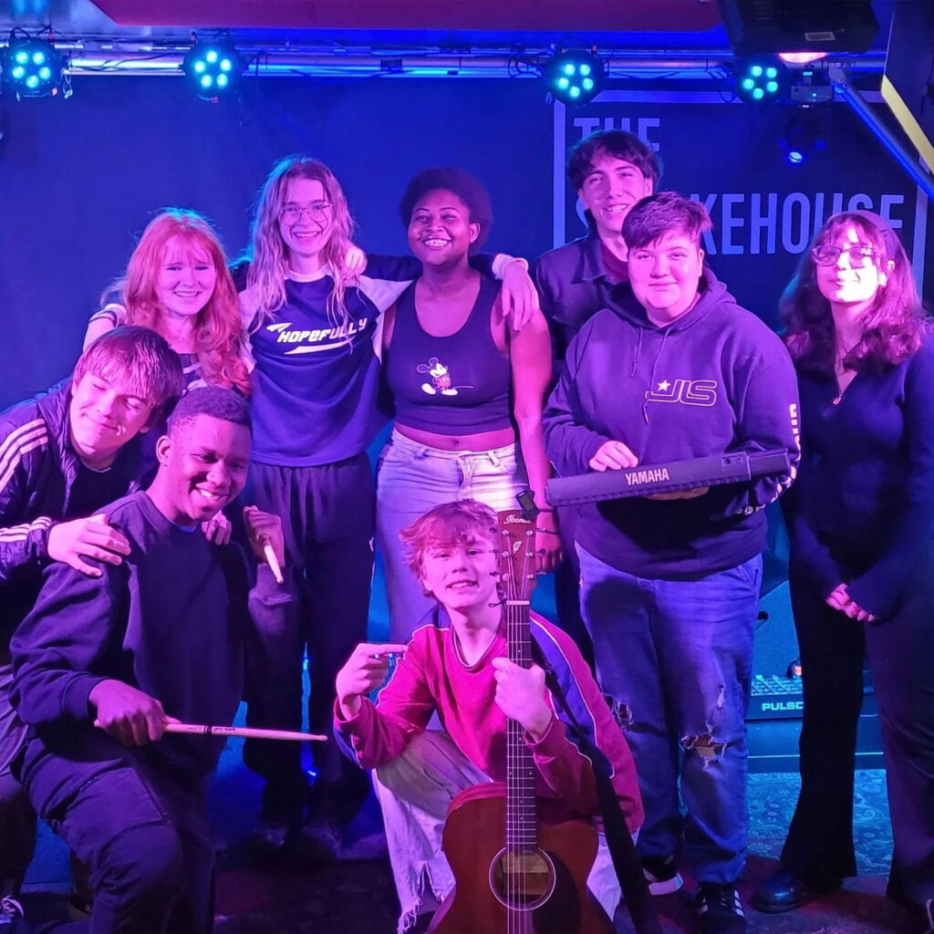 A group of nine young people pose on stage under colourful lights. One holds drumsticks, another holds a guitar, and another holds a keyboard. Everyone is smiling and standing in front of a sign that reads The Smokehouse.