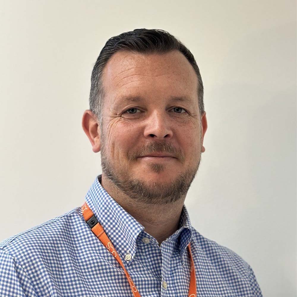 A man with short dark hair and a trimmed beard is wearing a blue checked shirt and an orange lanyard, standing against a plain light-coloured background, smiling slightly at the camera.