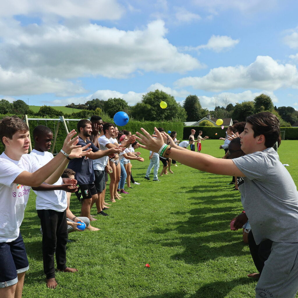 A group of children and teenagers stand in two queues on a grassy field, tossing water balloons to each other on a sunny day with scattered clouds. Some balloons are mid-air, and everyone is focused on catching them.