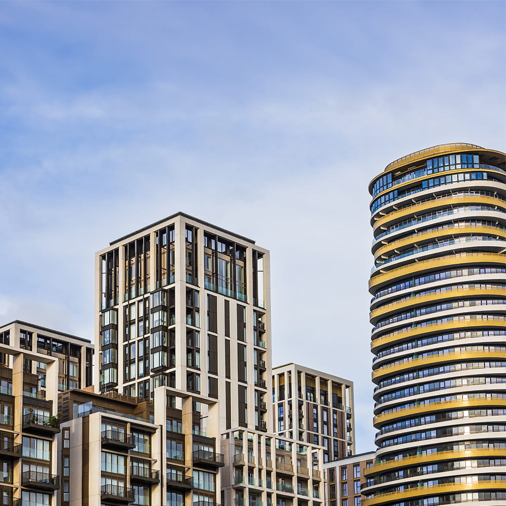 Modern high-rise block of flats with varied architectural designs against a blue sky with light clouds. The structures feature glass windows, balconies, and a mix of straight and curved lines.