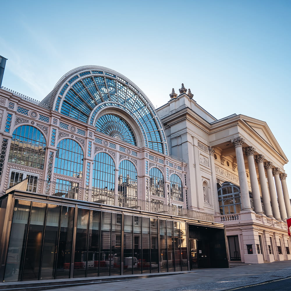 A grand building with an ornate glass arched façade and classical columns under a clear blue sky, seen from street level.