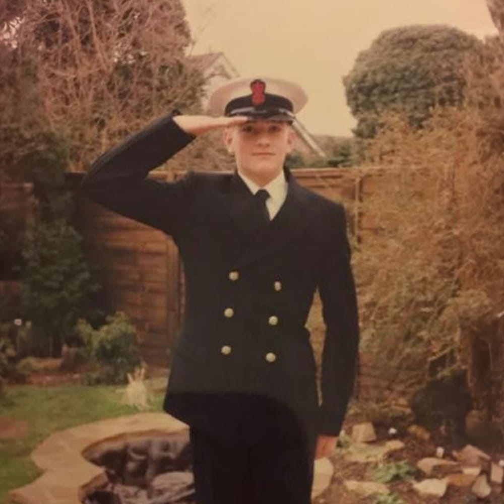 A person in a navy uniform with a white hat stands outdoors in a garden, saluting with their right hand. There are trees, shrubs, a wooden fence, and a small pond in the background.