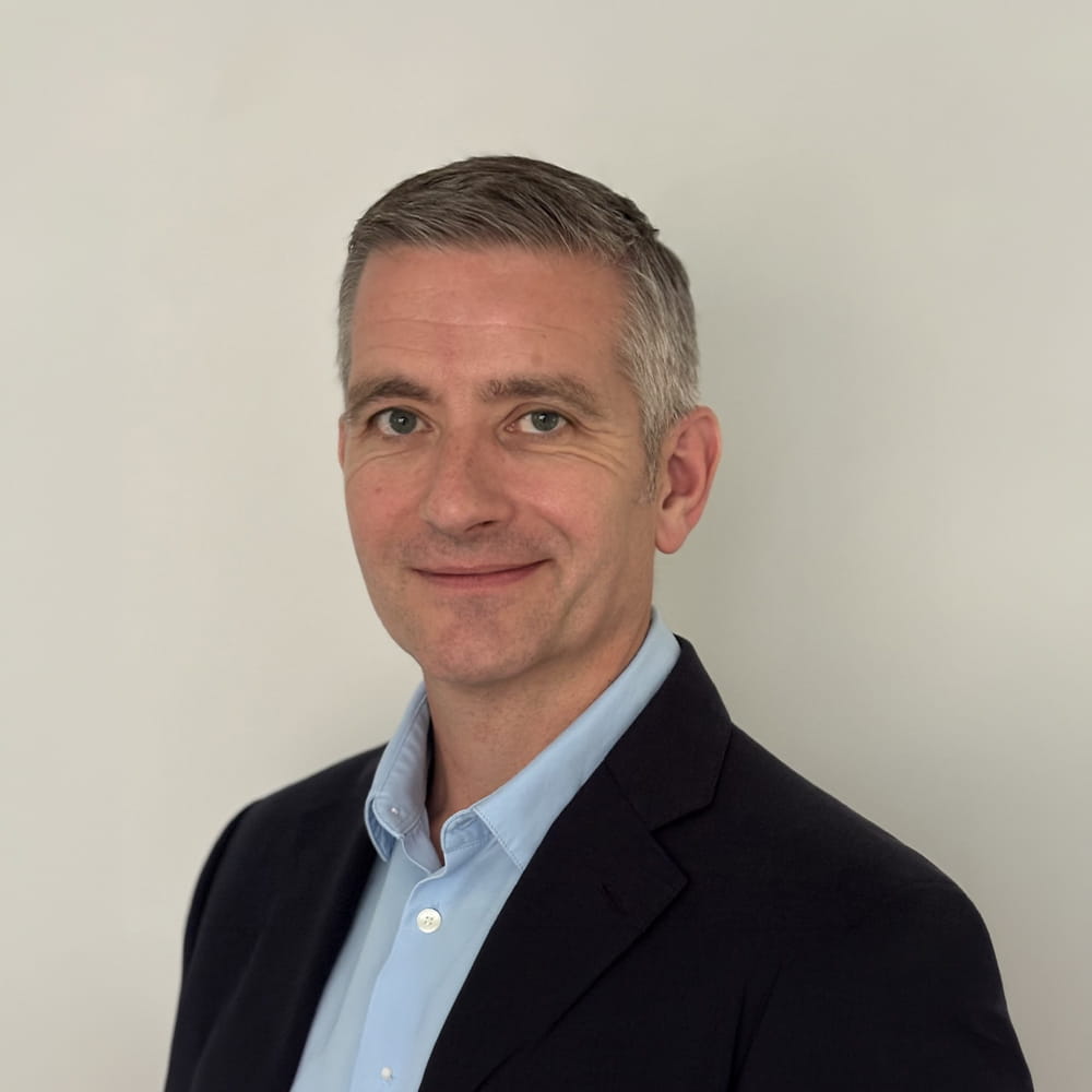 A man with short grey hair wearing a dark blazer and light blue shirt smiles slightly whilst posing in front of a plain, light-coloured background.