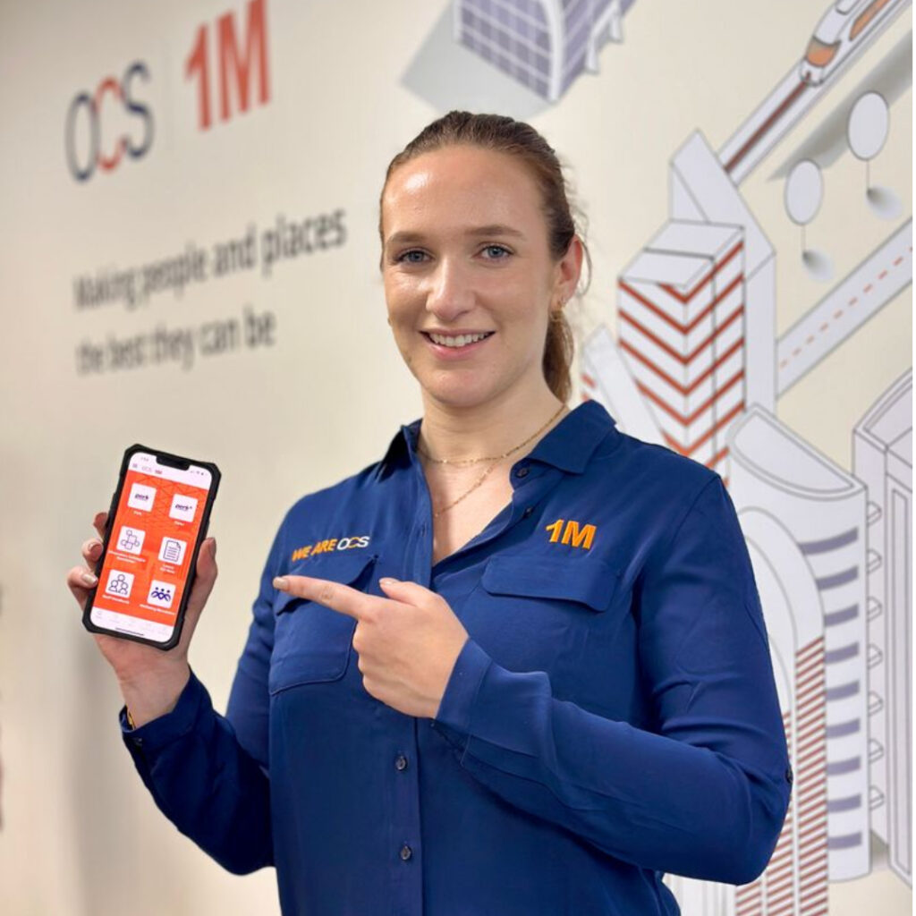 A woman in a blue shirt with 1M and WE ARE OCS embroidered holds up a mobile displaying an app with red icons. She stands in front of a wall with OCS branding and city illustrations.