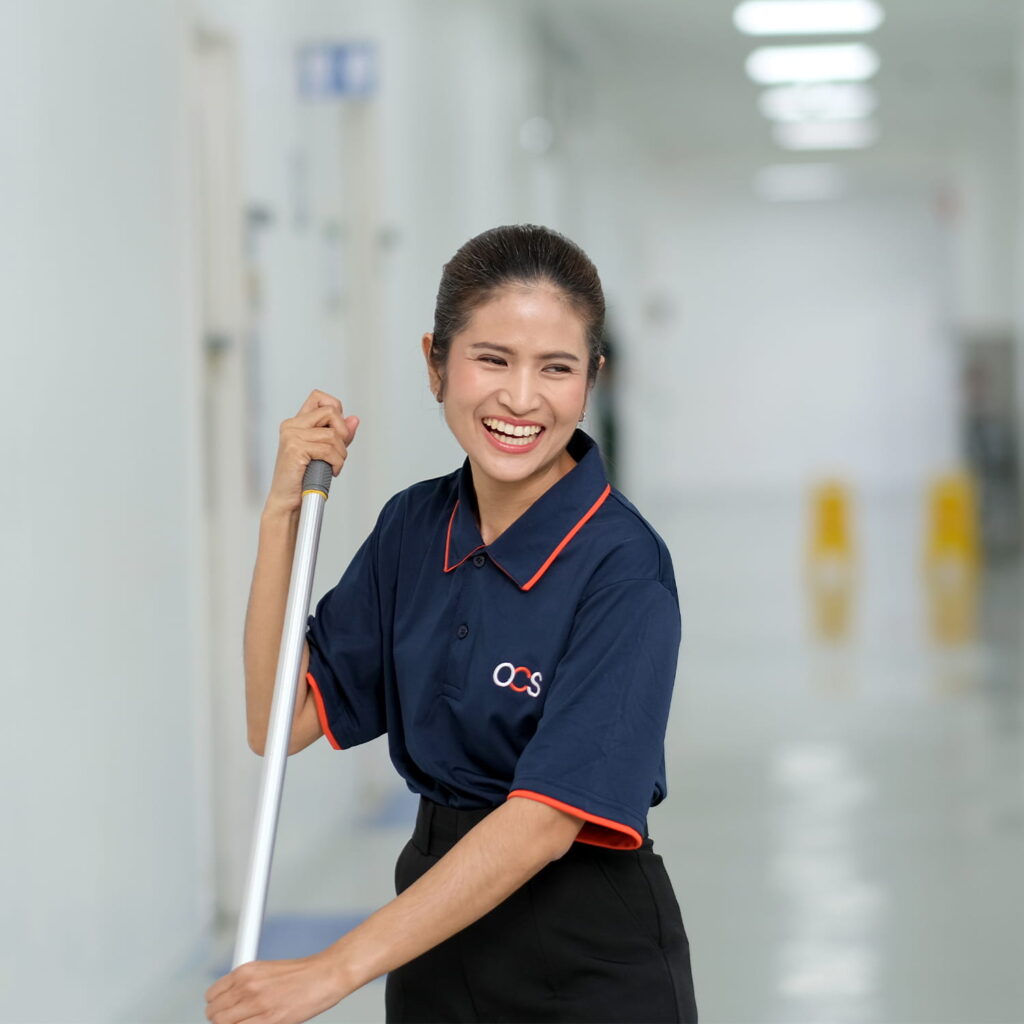 A smiling woman in a navy blue uniform with red trim is mopping a bright, clean hallway. She appears happy and focused on her work. The hallway is well-lit with blurred signage and caution signs in the background.