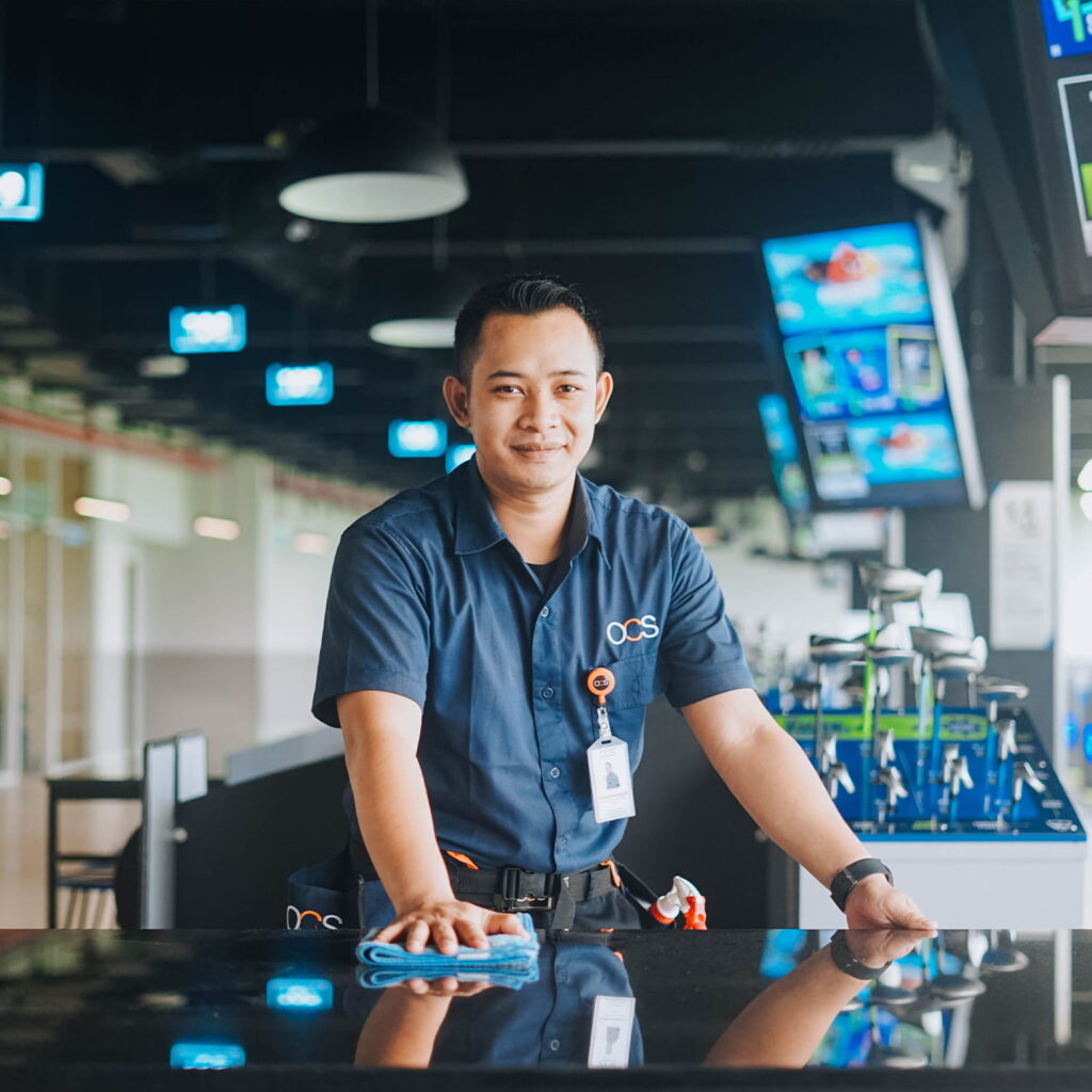 A smiling maintenance worker in a blue uniform wipes a countertop at an indoor facility, with golf clubs and multiple screens visible in the background.