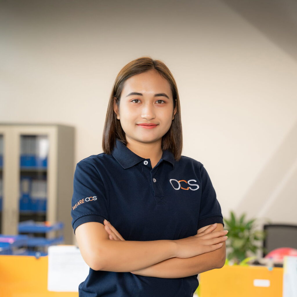 A woman with straight brown hair, wearing a navy blue collared OCS shirt, stands indoors with her arms crossed and smiles confidently at the camera. Office furniture and shelves are visible in the background.
