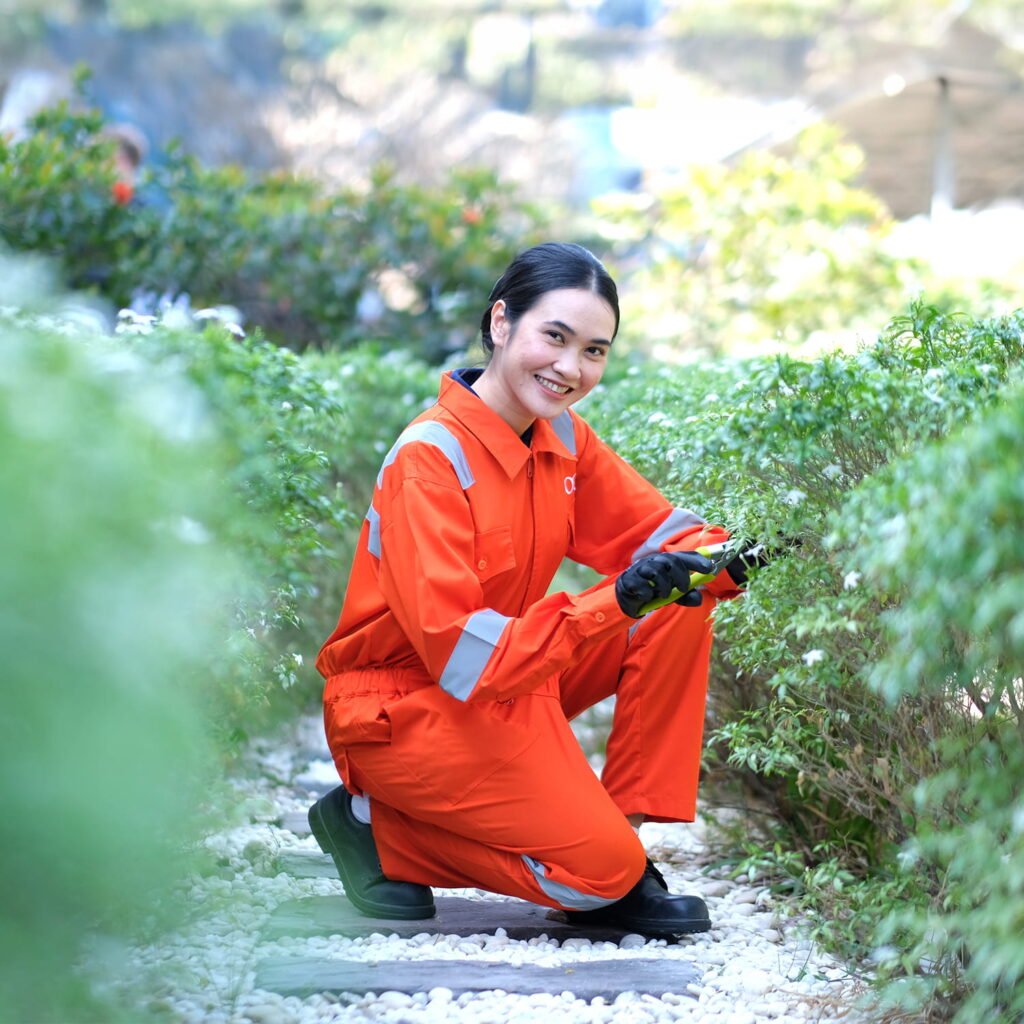 A woman wearing an orange work uniform and black gloves is crouching and trimming bushes in a garden. She is smiling at the camera and surrounded by greenery on a stone path.