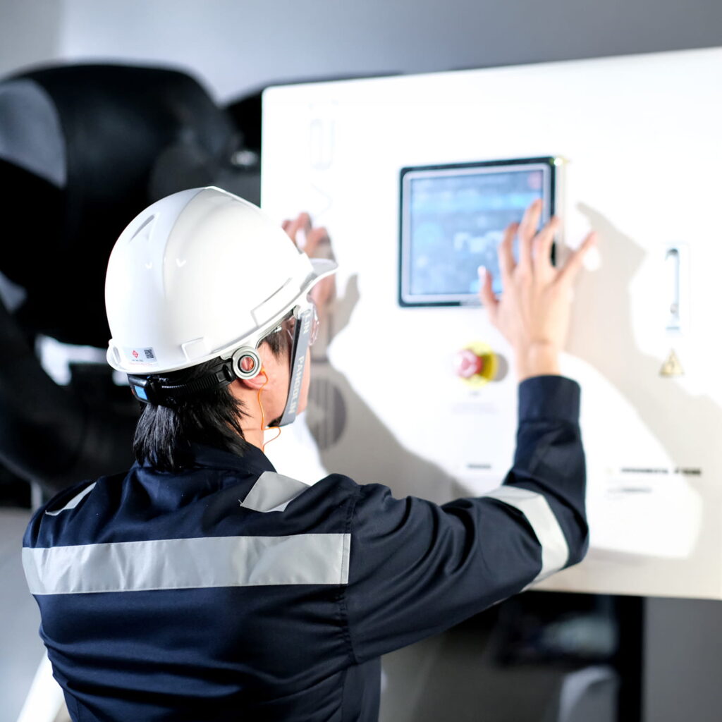 A person wearing a white hard hat, safety goggles, and a dark uniform operates a touchscreen control panel on industrial machinery.