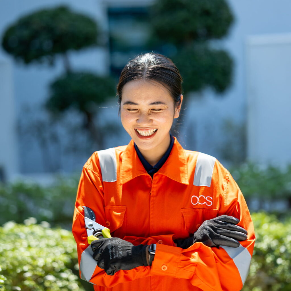 A smiling person wearing an orange work uniform with reflective stripes and black gloves stands outdoors, holding pruning shears with folded arms. Greenery and blurred trees appear in the background.