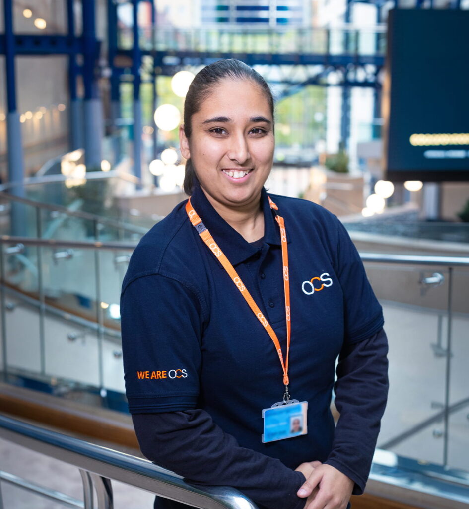 A smiling person with dark hair tied back wears an OCS uniform and an orange lanyard with an ID badge, standing indoors by a metal railing in a modern building.