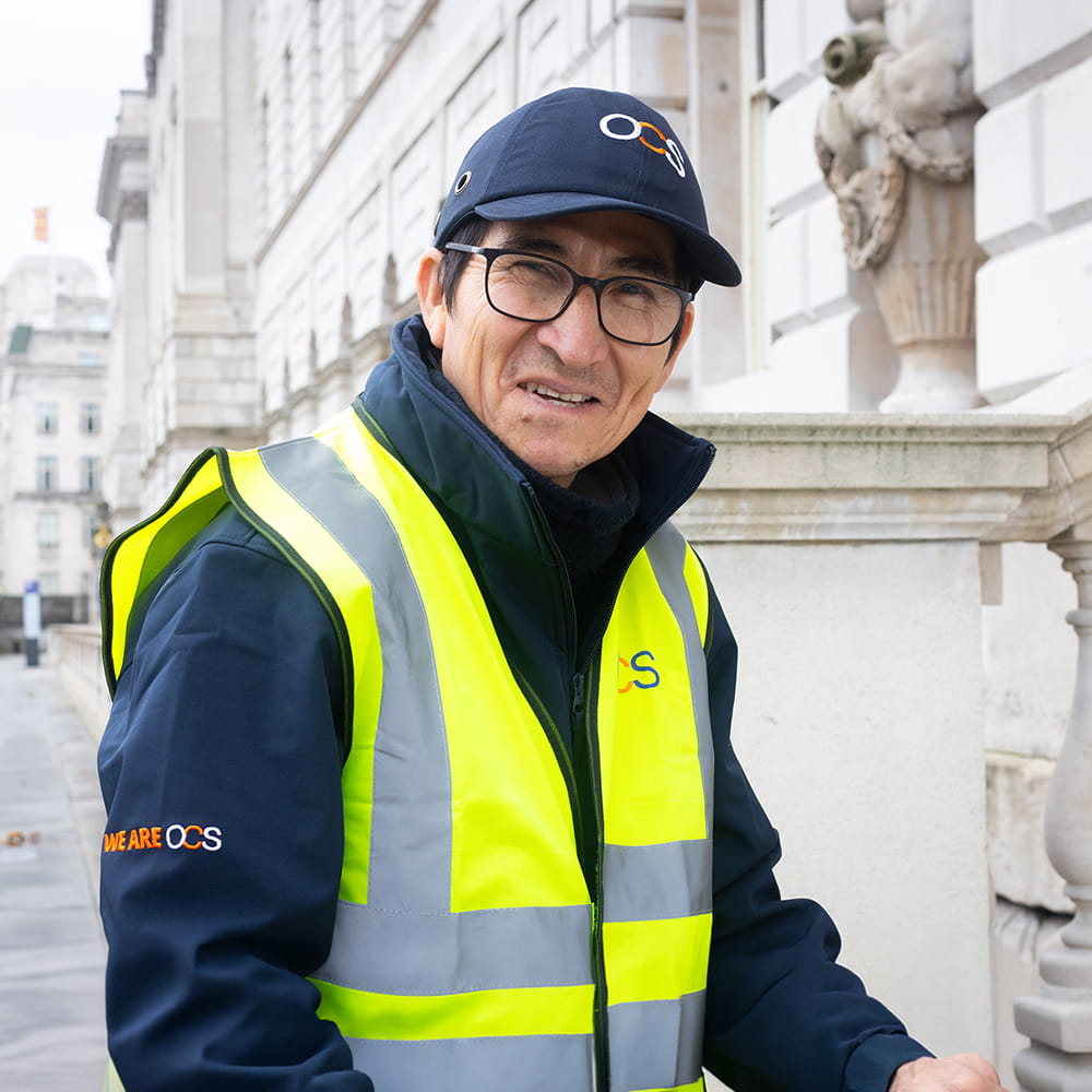 A man wearing glasses, a navy OCS cap, and a high-visibility yellow safety vest stands smiling on a city pavement in front of a stone building with classical architectural details.