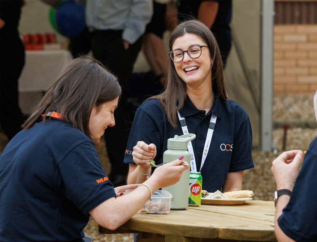 Two women in matching navy shirts sit at an outdoor table, smiling and laughing whilst eating lunch. A can of 7UP and a water bottle are visible. Other people are blurred in the background.