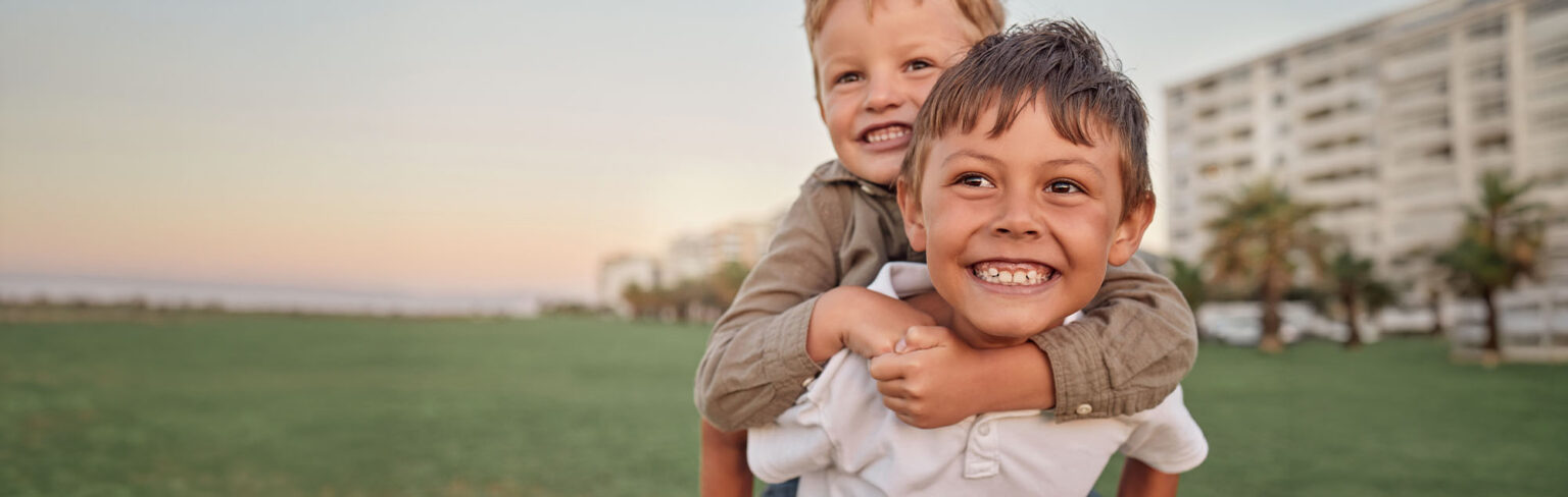 Two smiling young boys play outside; one boy gives the other a piggyback. They are on a grassy field with tall buildings and palm trees in the background at sunset.