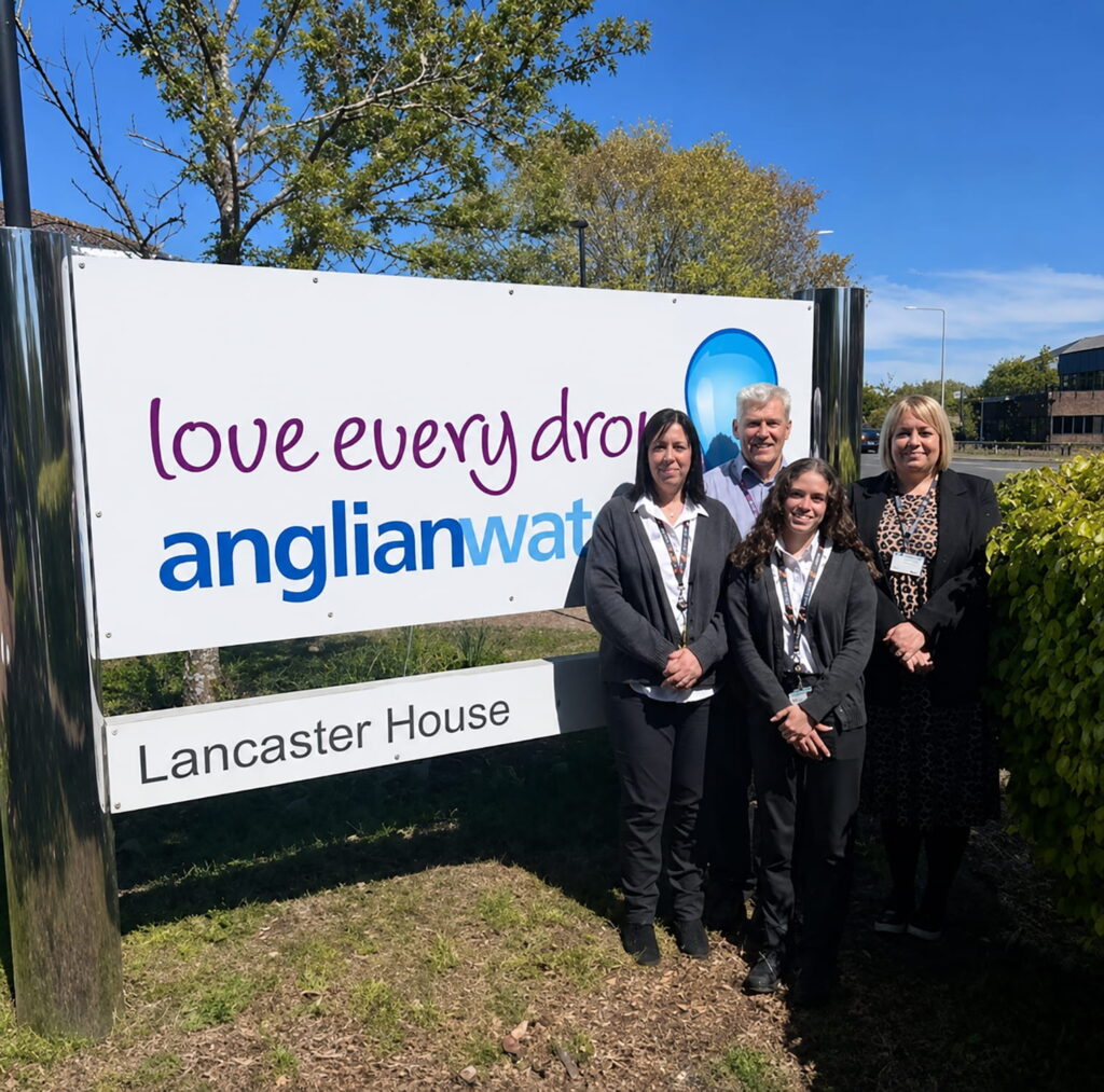 Four people stand smiling in front of an Anglian Water sign that reads love every drop and Lancaster House on a sunny day, with greenery and trees in the background.