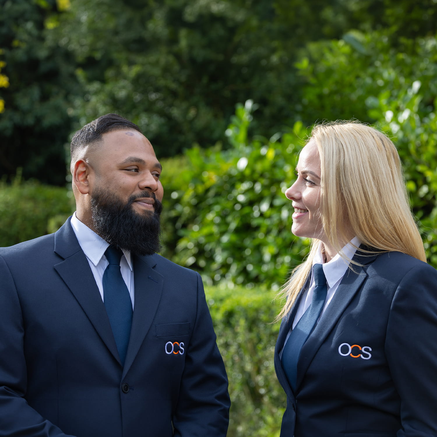 Two people in matching dark suits with OCS logos, standing outdoors and smiling at each other, with greenery and trees in the background.