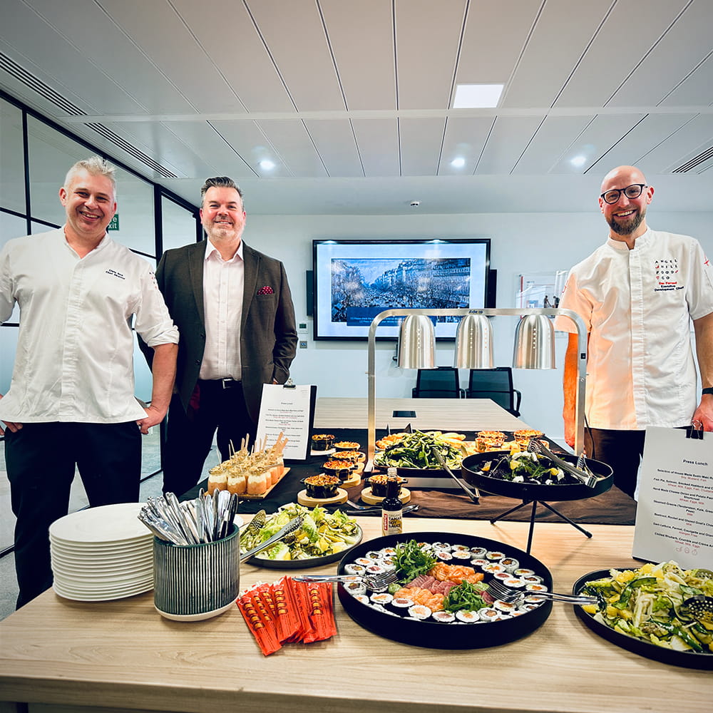Three men stand behind a table filled with various plated dishes, including sushi, salads, and starters, in a modern, well-lit office setting. Plates, cutlery, and menus are arranged at the front of the table.