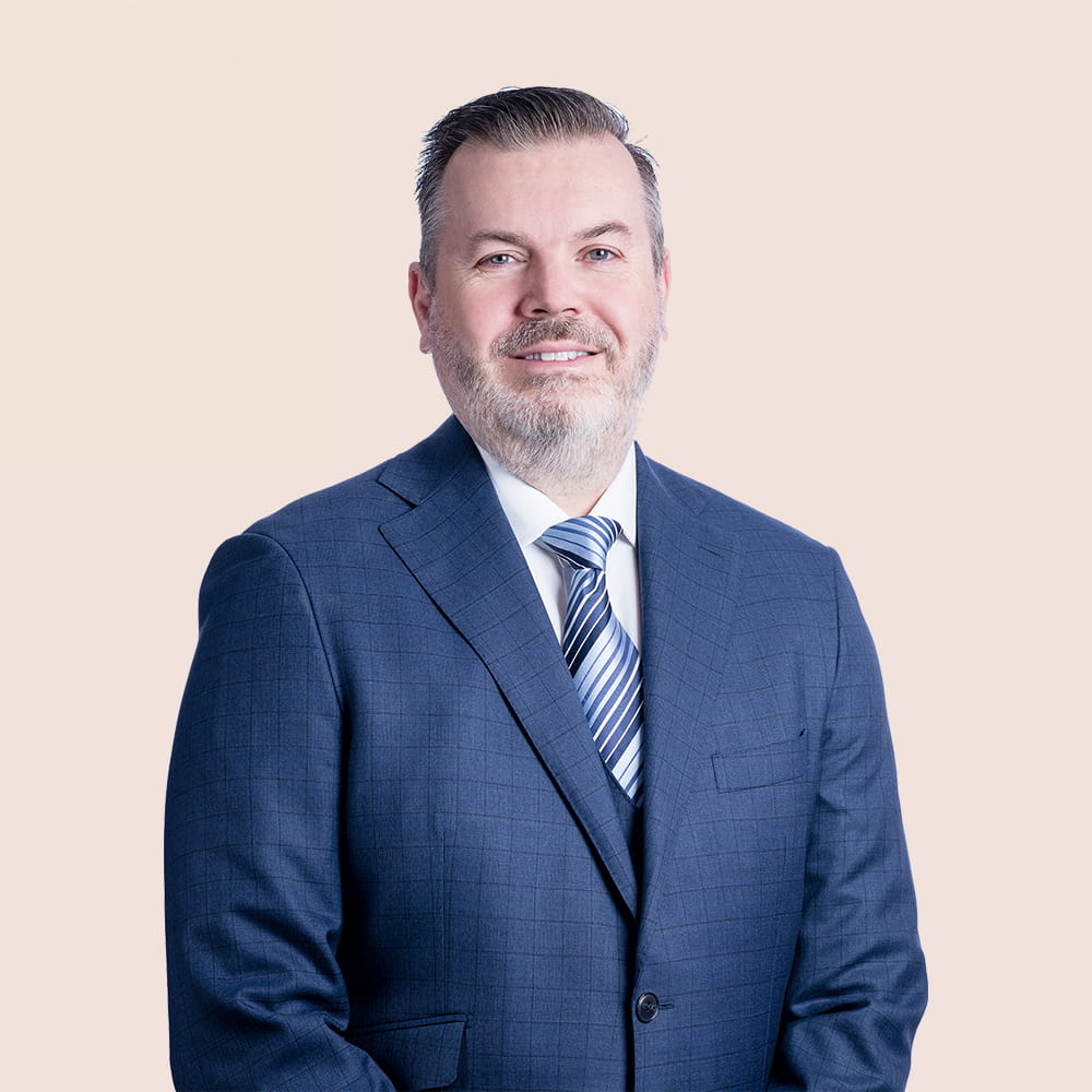 A man in a blue checked suit, white shirt, and striped tie is smiling and facing the camera against a plain light beige background.