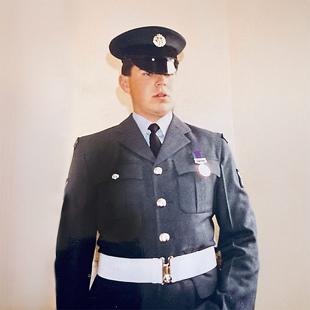 A man in a formal military uniform with a cap, dark jacket, light blue shirt, and white belt stands indoors against a plain light-coloured wall. He wears medals on his chest and looks slightly to the side.