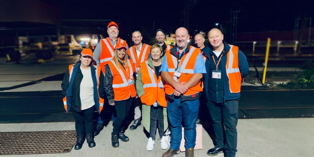 A group of ten people wearing orange safety vests and casual clothing stand together outdoors at night, smiling at the camera. Construction or railway equipment is visible in the background.